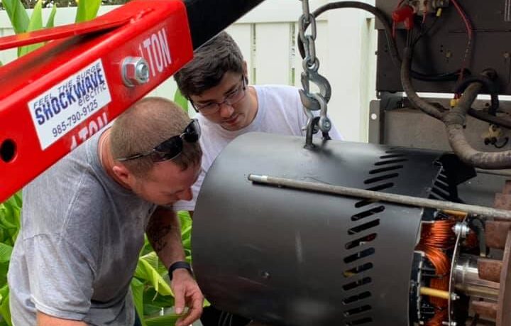Two individuals working on the maintenance of a large electrical motor or generator, using a red engine hoist for assistance.